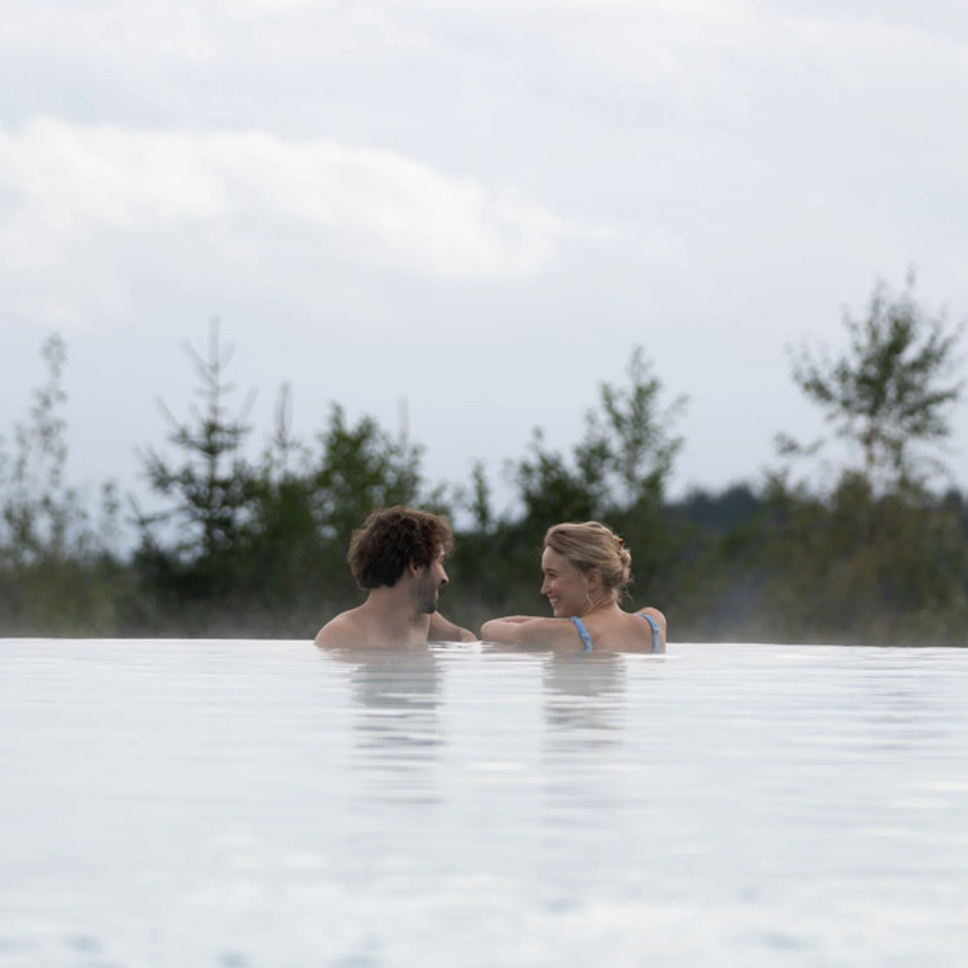 Paar entspannt sich im Infinity-Pool mit Wald im Hintergrund bei bewölktem Himmel