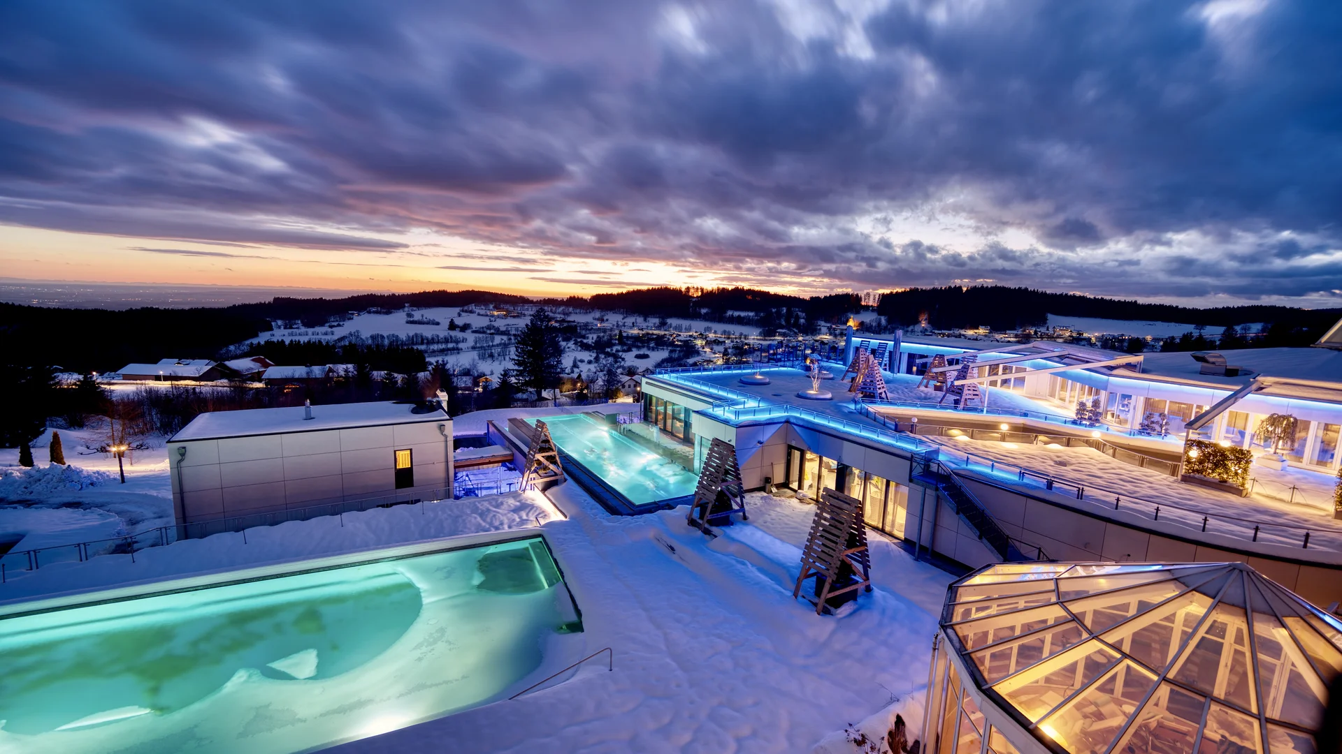Moderner Spa mit beleuchteten Pools bei Sonnenuntergang in verschneiter Landschaft