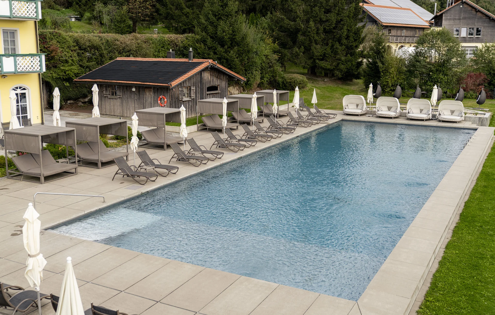 Großer Pool mit Liegestühlen und Holzhütte vor Waldkulisse