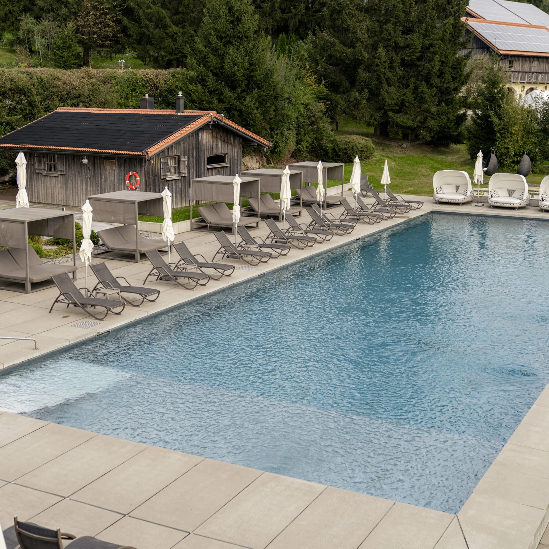 Großer Pool mit Liegestühlen und Holzhütte vor Waldkulisse