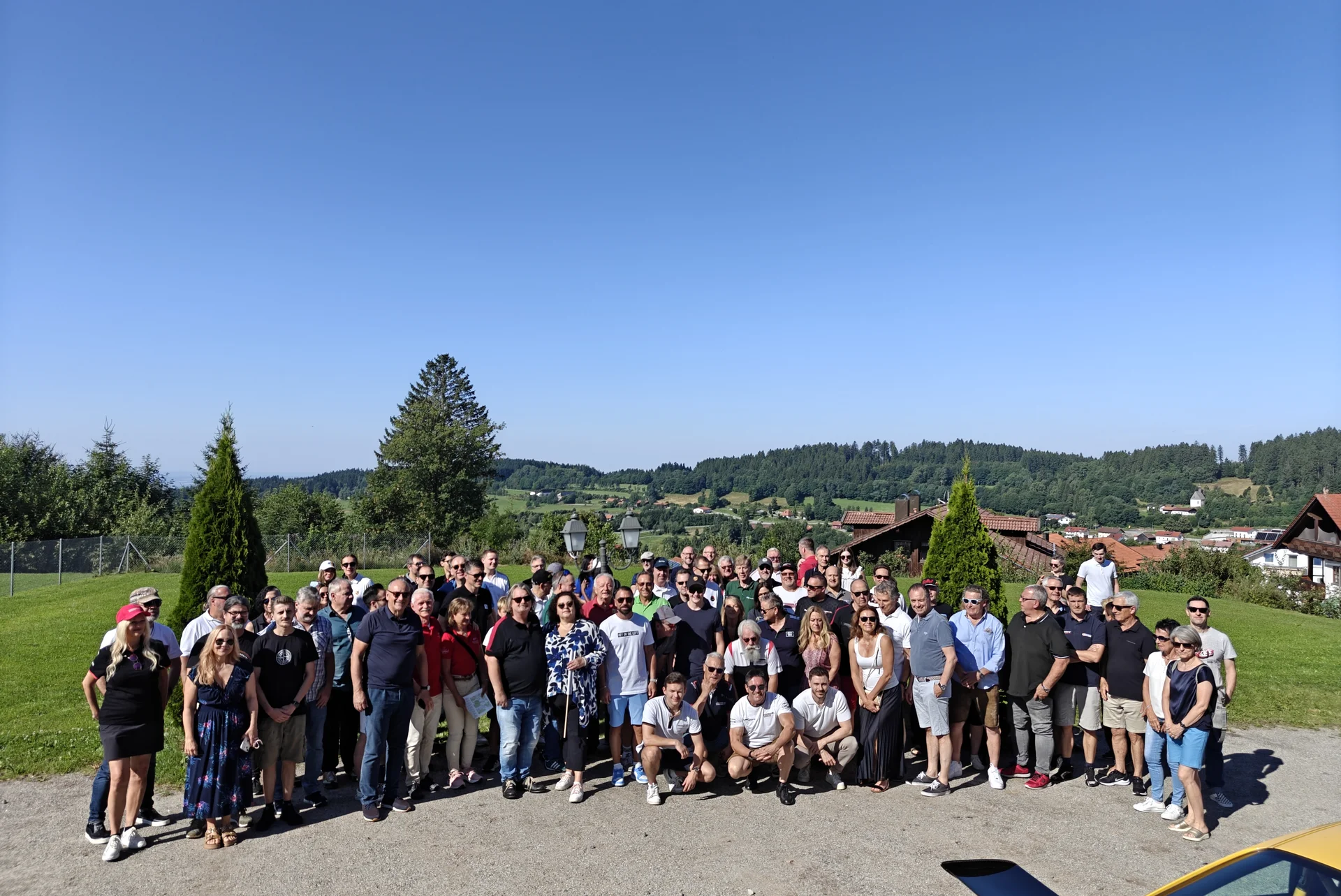 Große Gruppe von Menschen draußen vor einer ländlichen Kulisse bei klarem Himmel.