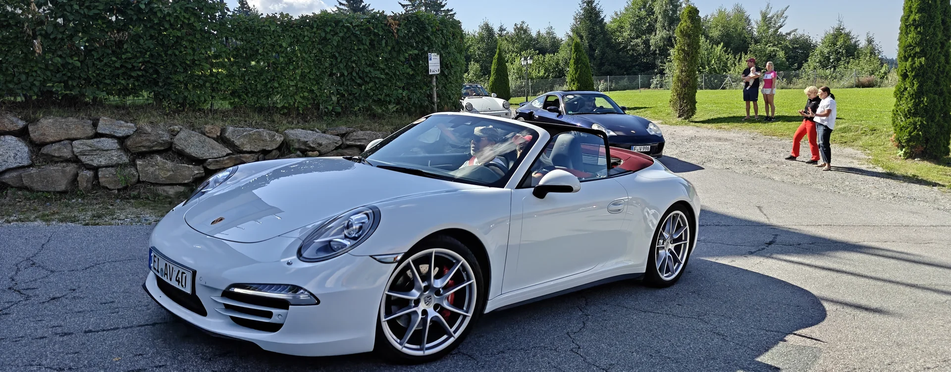 Weiße Porsche Cabrio auf Straße bei sonnigem Wetter mit Menschen und Bäumen
