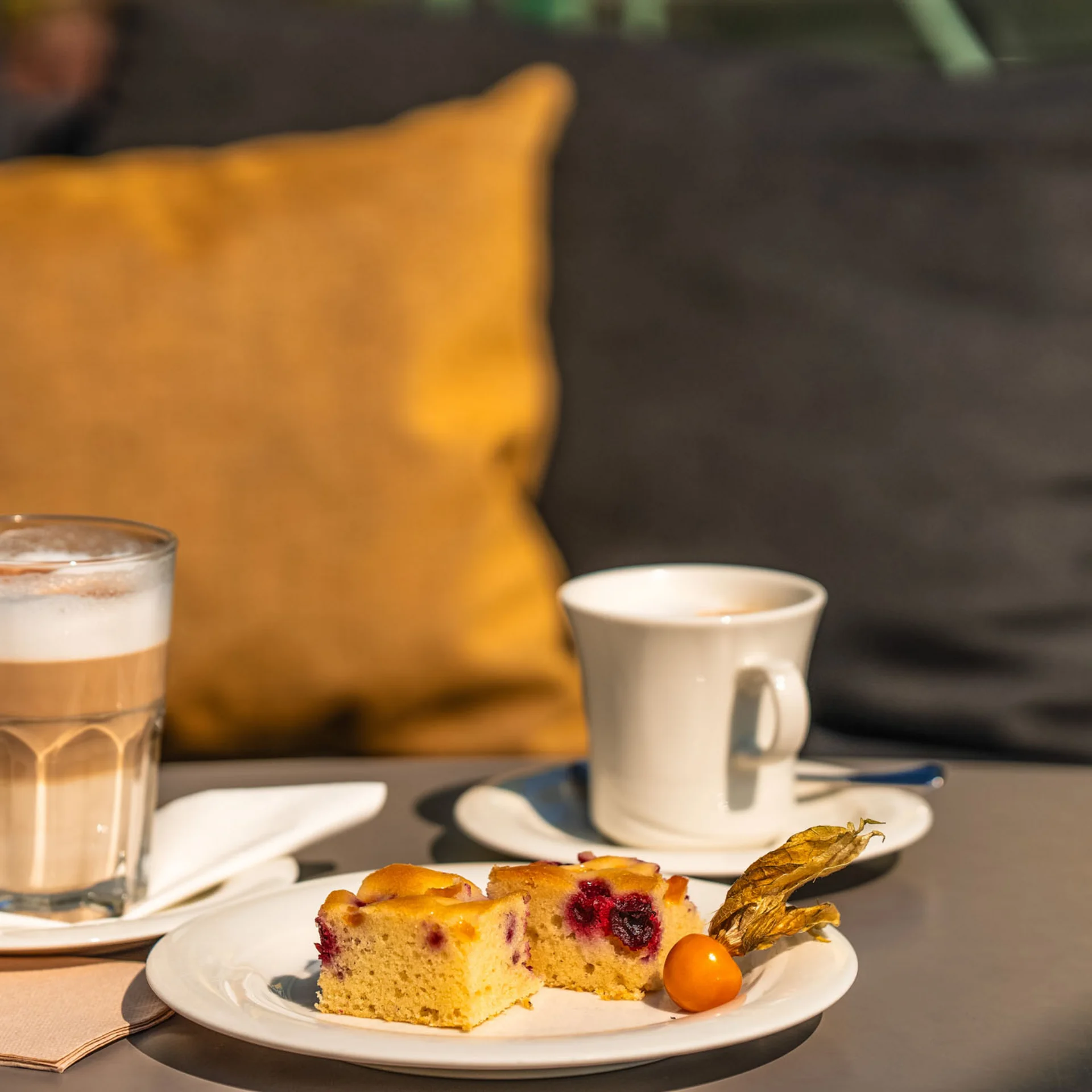 Kuchenstück und zwei Kaffeegetränke auf einem Tisch vor gelbem Kissen