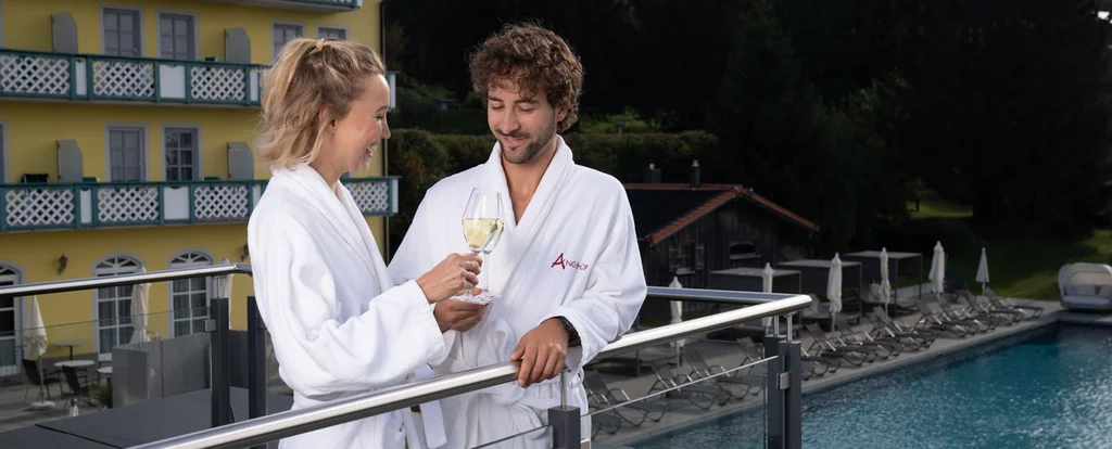 Paar in weißen Bademänteln trinkt Sekt auf Balkon mit Pool und Tannen im Hintergrund