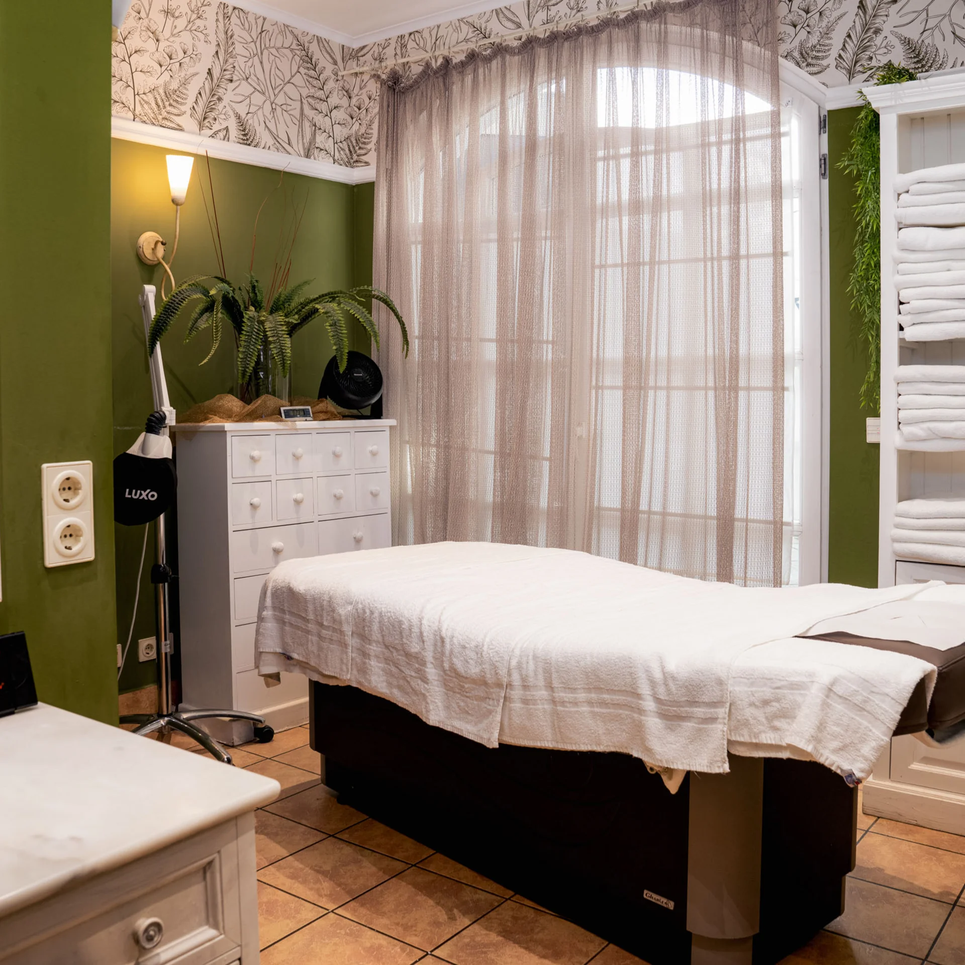 Behandlungsraum mit Massageliege, grünen Wänden und Regalen voller weißer Handtücher