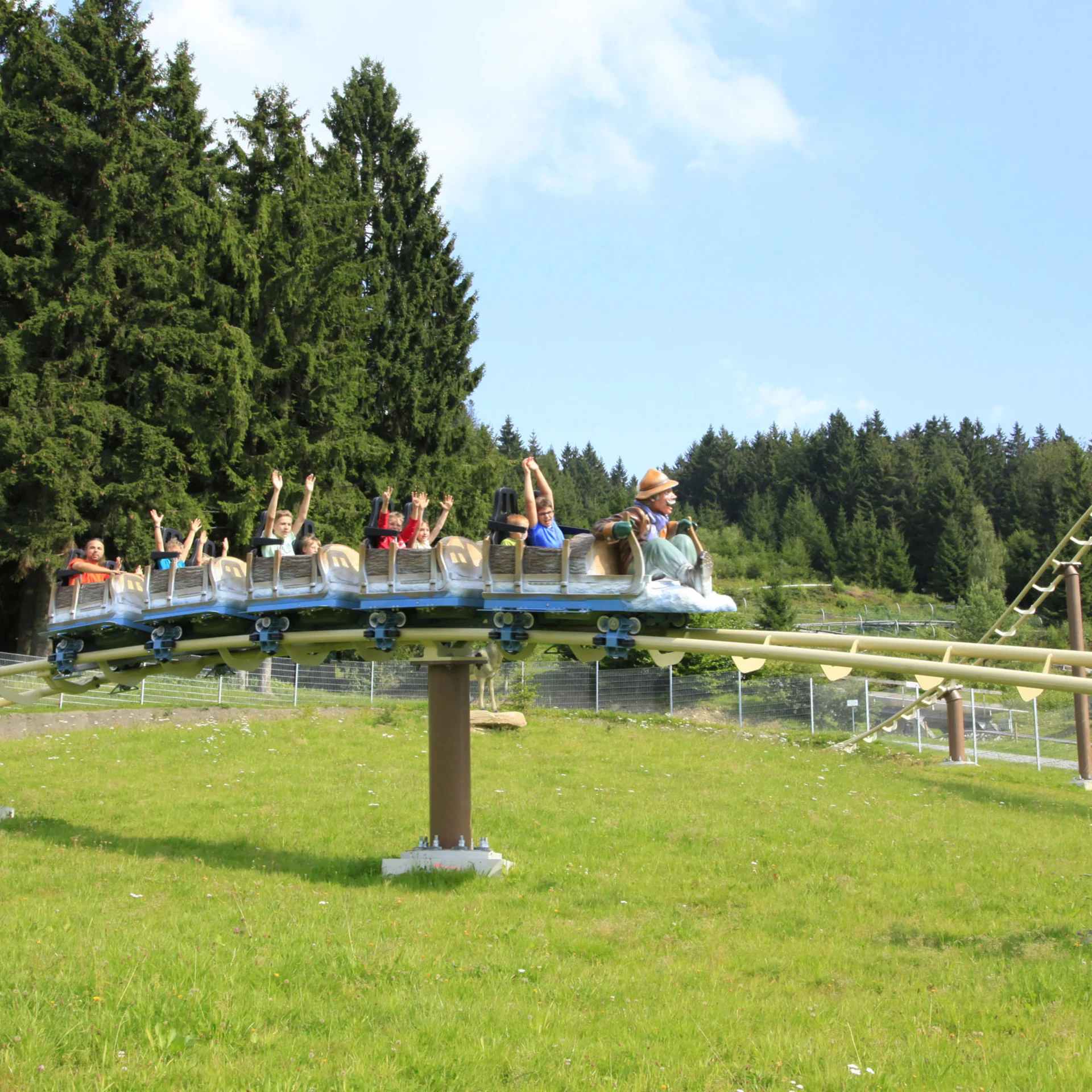 Menschen fahren eine Achterbahn in einer grünen Berglandschaft bei Sonnenschein