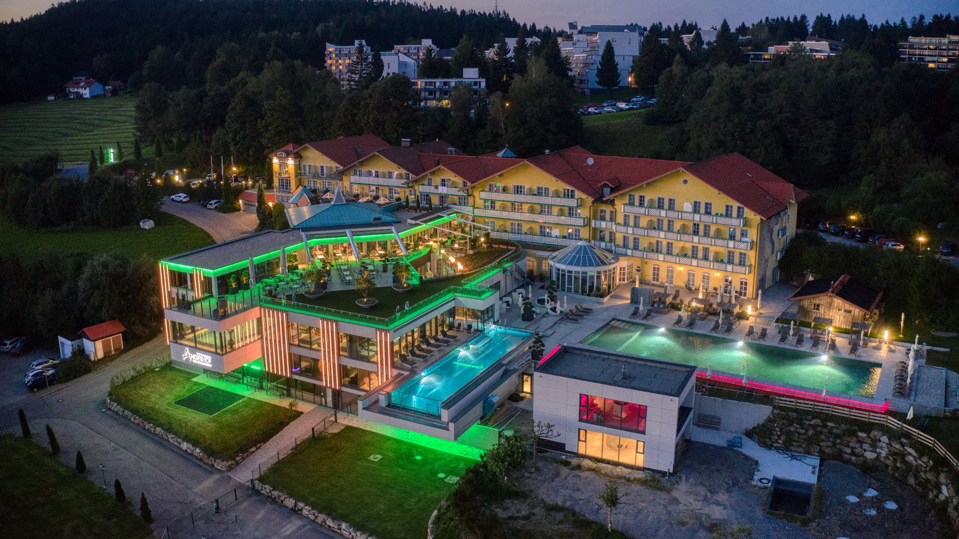 Angerhof: Urlaub in Panoramalage im Bayerischen Wald Modernes Hotel mit beleuchtetem Pool und umliegenden Gebäuden bei Abenddämmerung