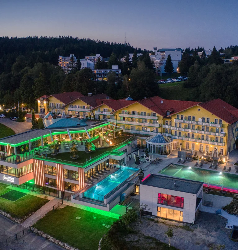 Die Adresse für Wellness in Bayern: Ihr Hotel in St. Englmar Modernes Hotel mit beleuchtetem Pool und umliegenden Gebäuden bei Abenddämmerung