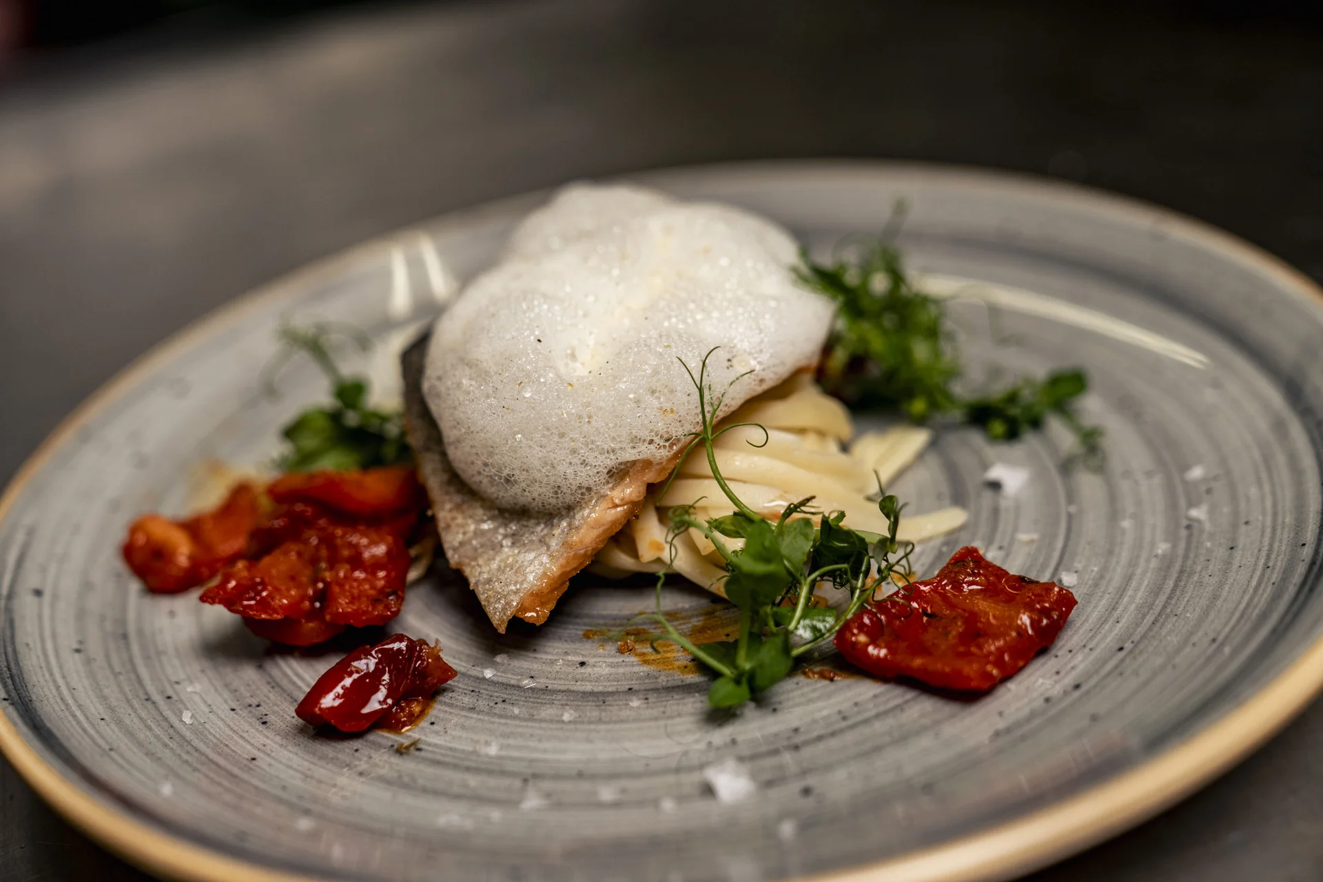 Gourmetgericht mit Schaum, Kräutern und getrockneten Tomaten auf grauem Teller