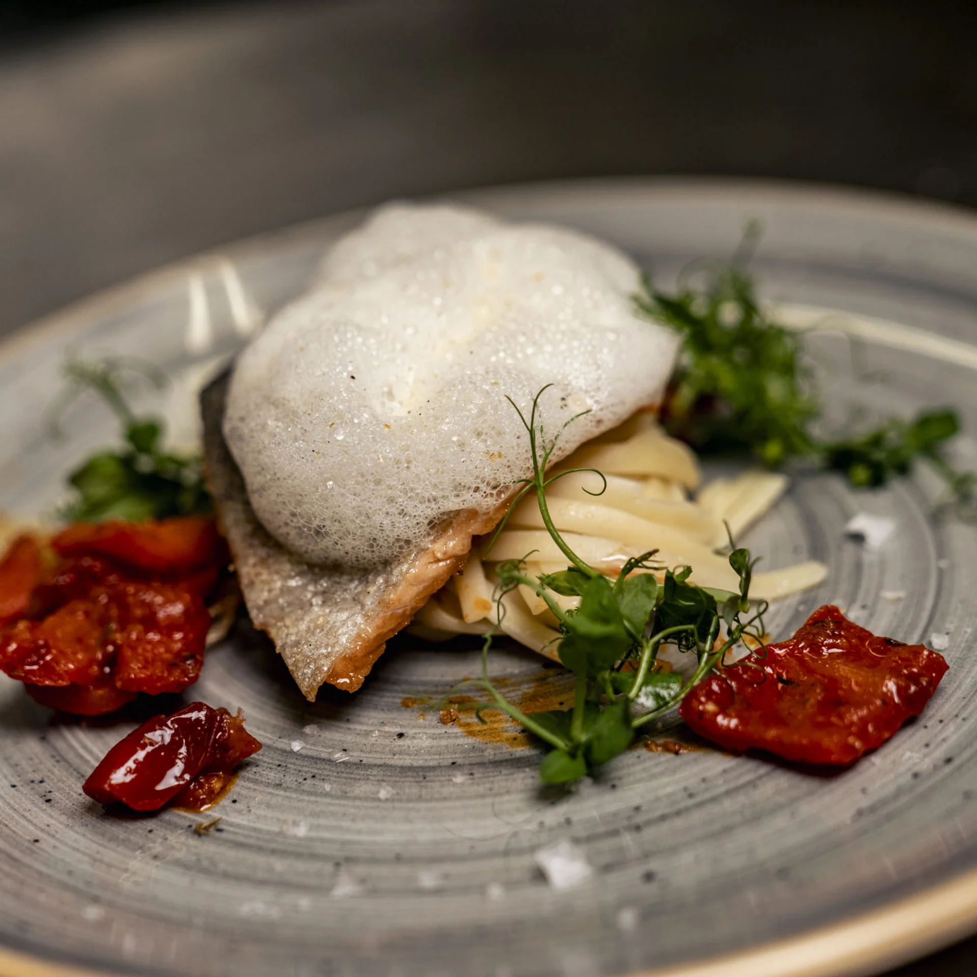Gourmetgericht mit Schaum, Kräutern und getrockneten Tomaten auf grauem Teller