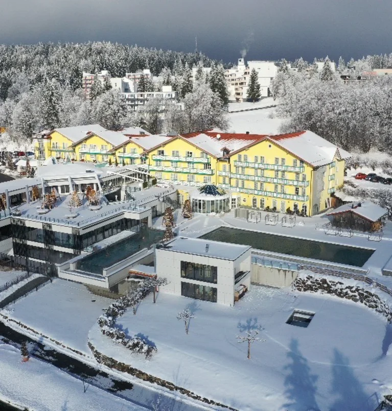 Verschneite Hotelanlage mit gelben Gebäuden und Außenpool, umgeben von winterlichen Bäumen