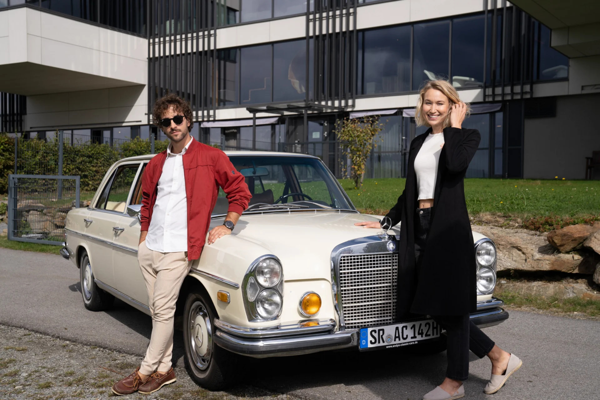 Mann und Frau lehnen an einem weißen Oldtimer vor einem modernen Gebäude