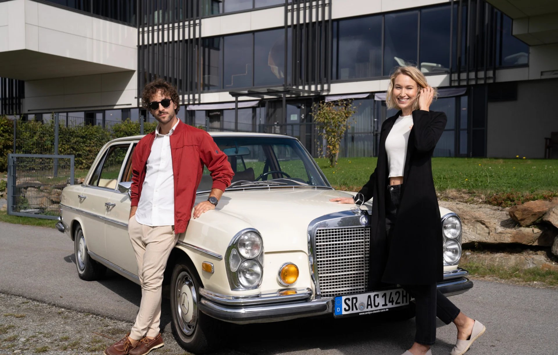 Mann und Frau lehnen an einem weißen Oldtimer vor einem modernen Gebäude