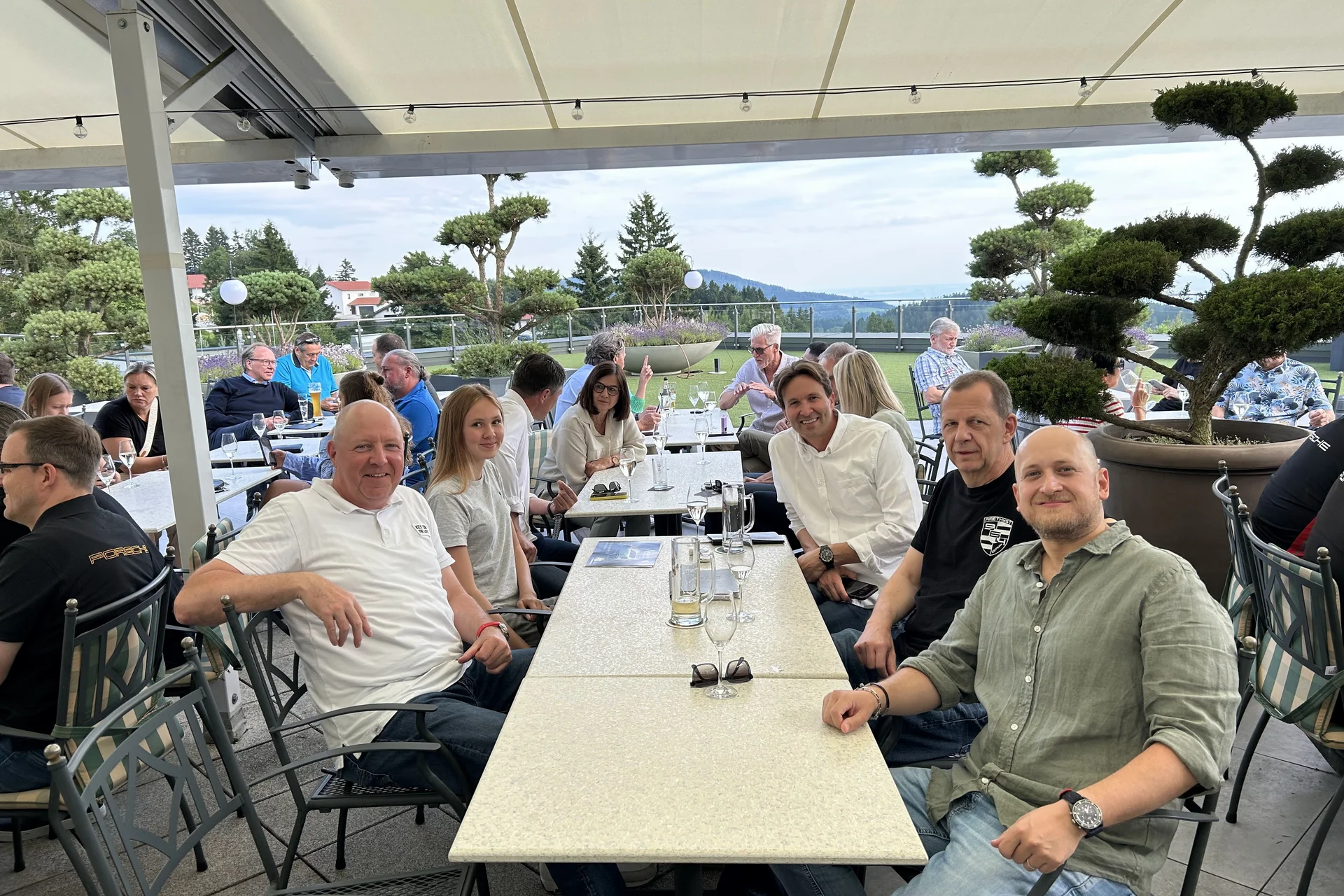 Menschen sitzen draußen an Tischen unter einem Sonnendach mit Bergblick