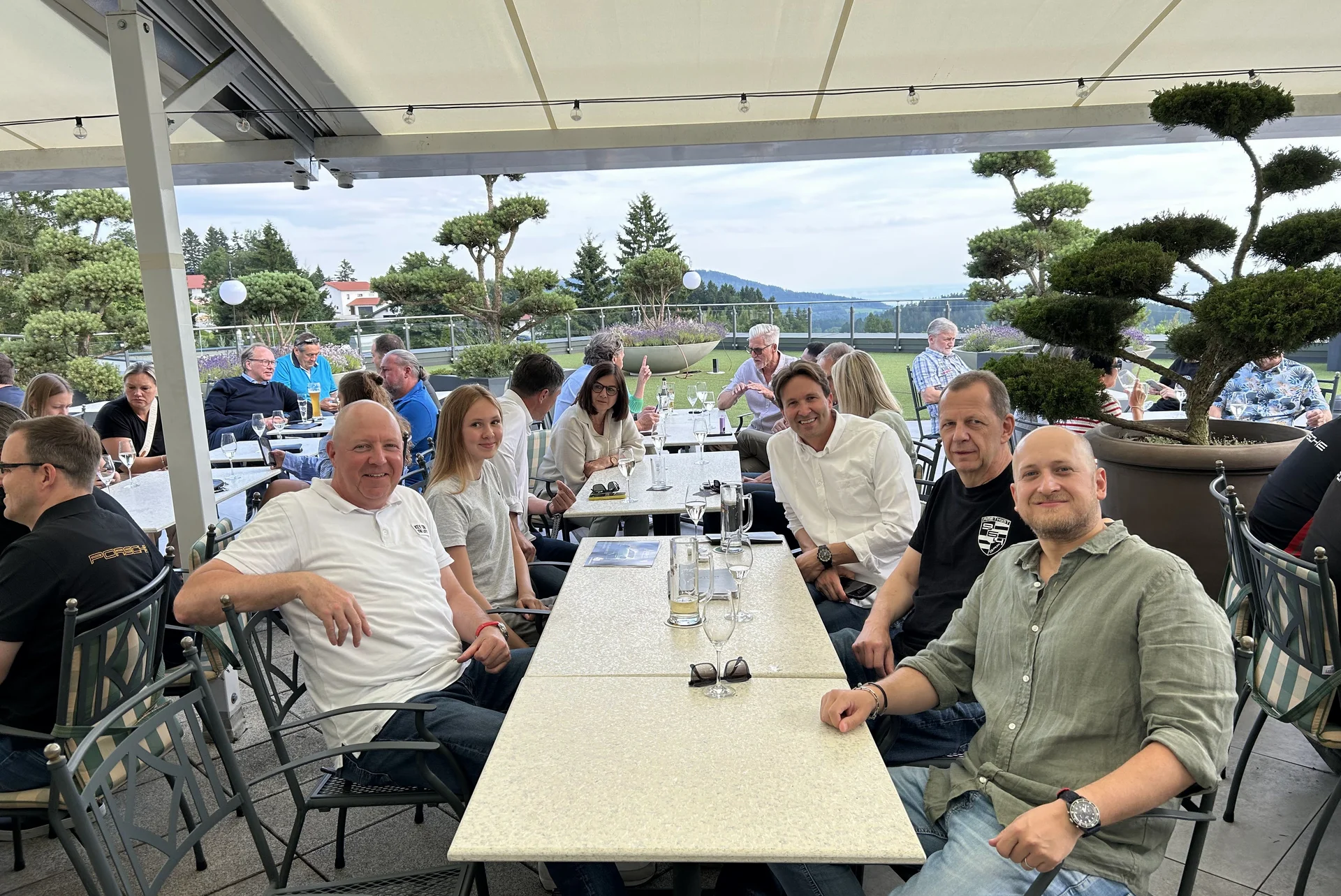 Menschen sitzen draußen an Tischen unter einem Sonnendach mit Bergblick