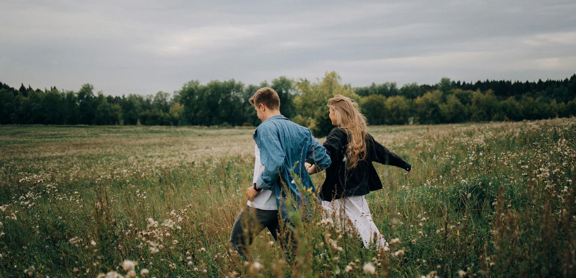 Paar hält Händchen und läuft durch ein Feld mit Bäumen im Hintergrund