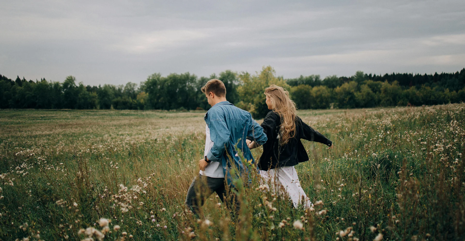 Paar hält Händchen und läuft durch ein Feld mit Bäumen im Hintergrund