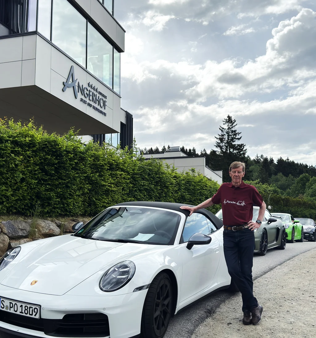 Mann lehnt an weißem Porsche vor Angerhof Hotel, weitere Autos und Bäume im Hintergrund