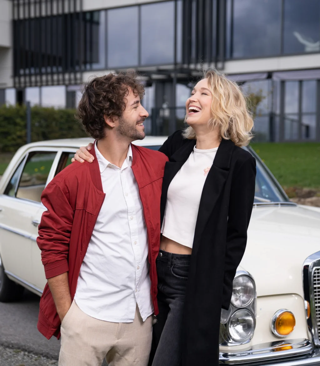 Lachendes Paar steht vor einem weißen Oldtimer-Mercedes vor modernem Gebäude