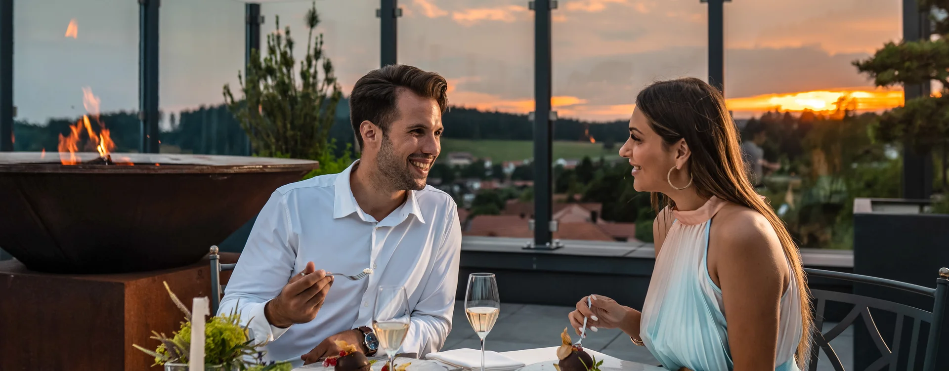 Paar beim Essen und Trinken auf einer Terrasse mit Sonnenuntergang im Hintergrund