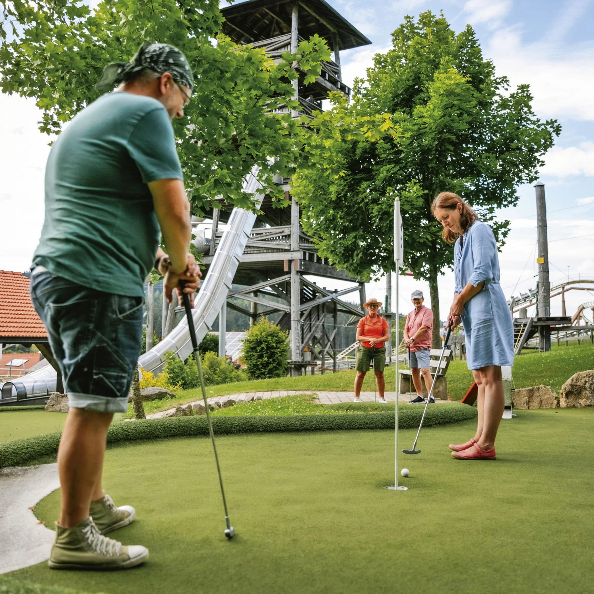 Personen spielen Minigolf auf einem Grün neben einem Freizeitpark mit Rutschen