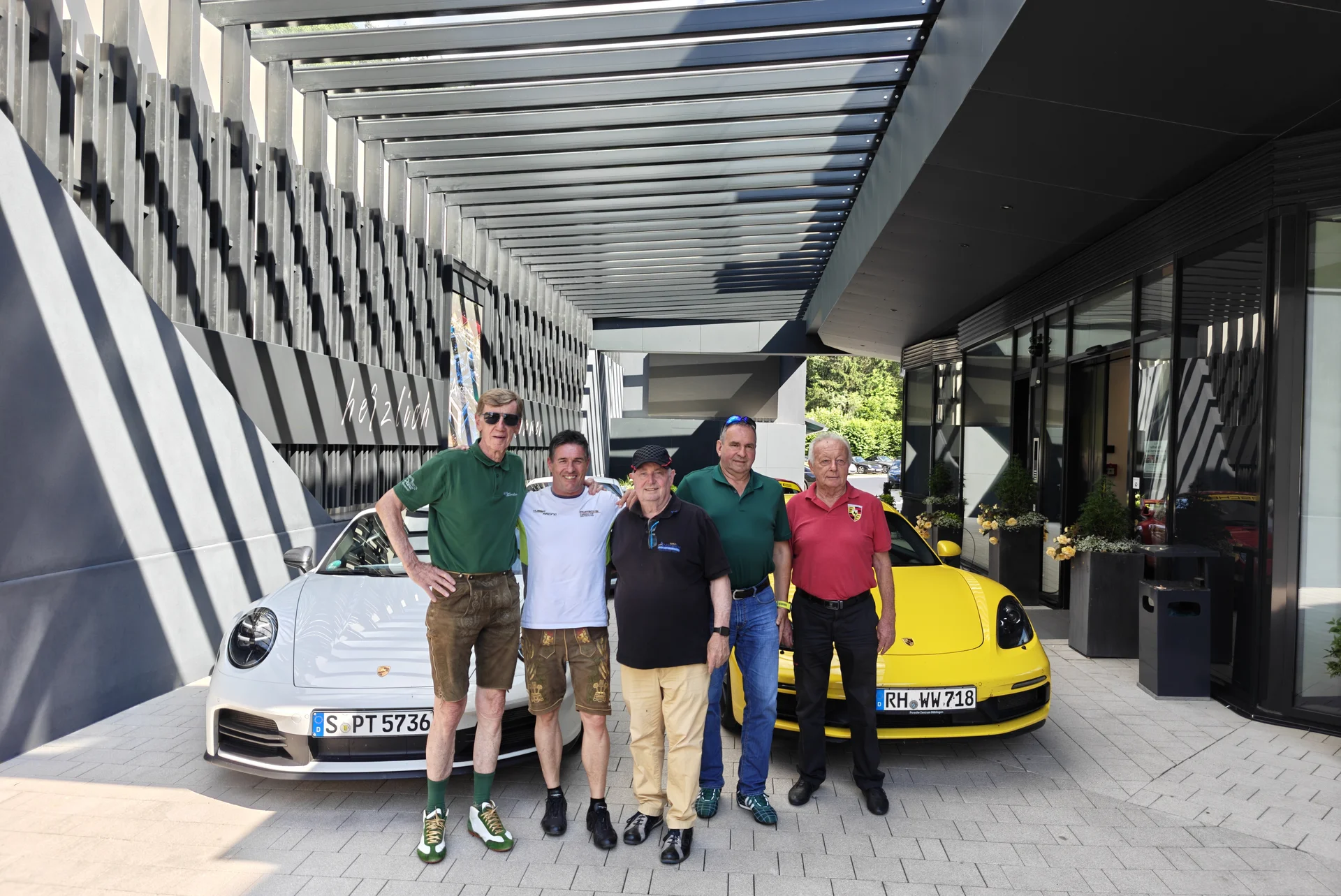 Fünf Männer posieren vor weißen und gelben Porsche Autos unter einem modernen Vordach.
