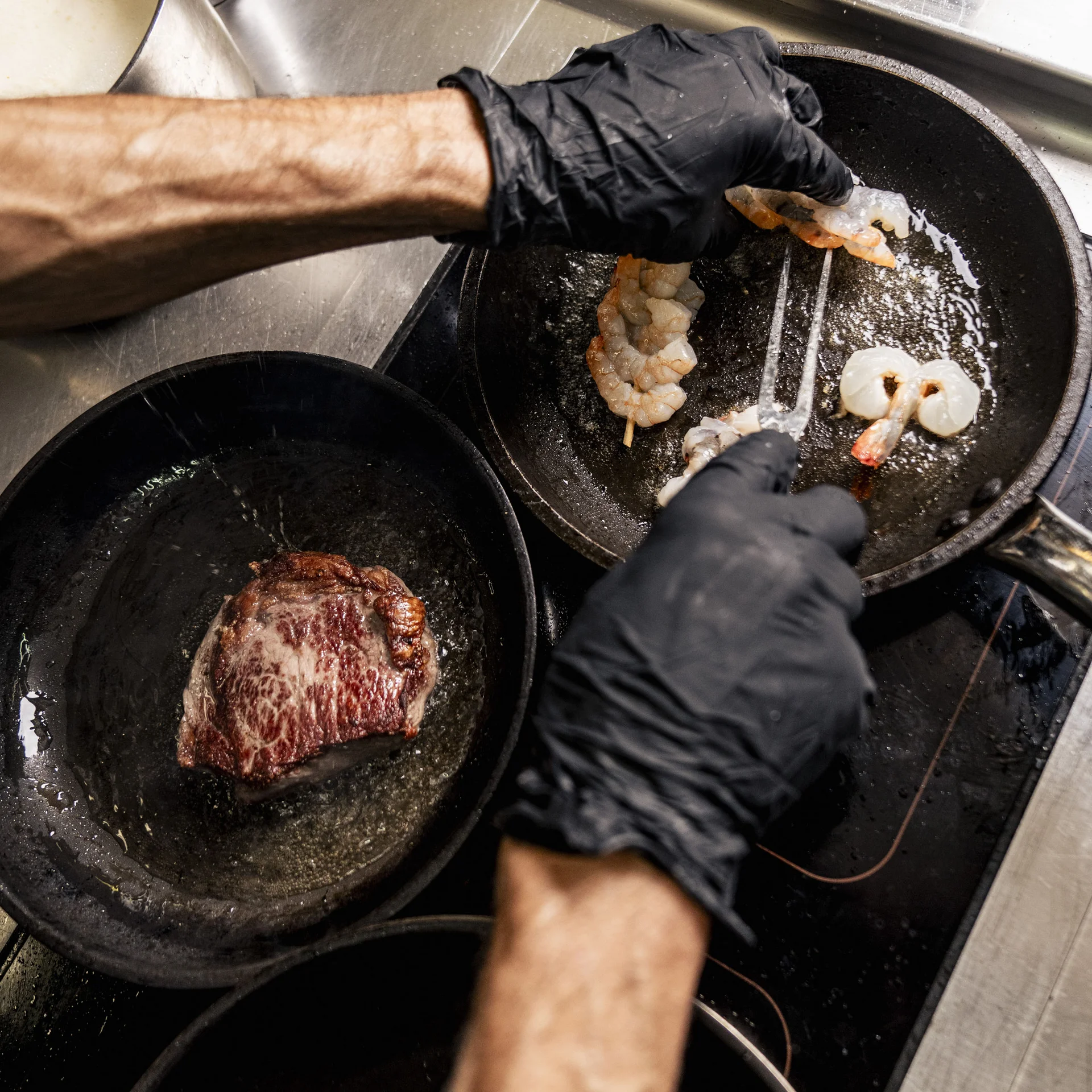 Person brät Garnelen und ein Steak in einer Pfanne mit schwarzen Handschuhen