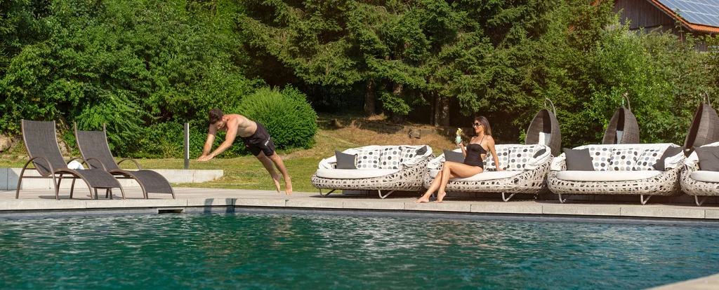 Mann springt ins Schwimmbecken, Frau sitzt entspannt mit Getränk auf Gartenliege