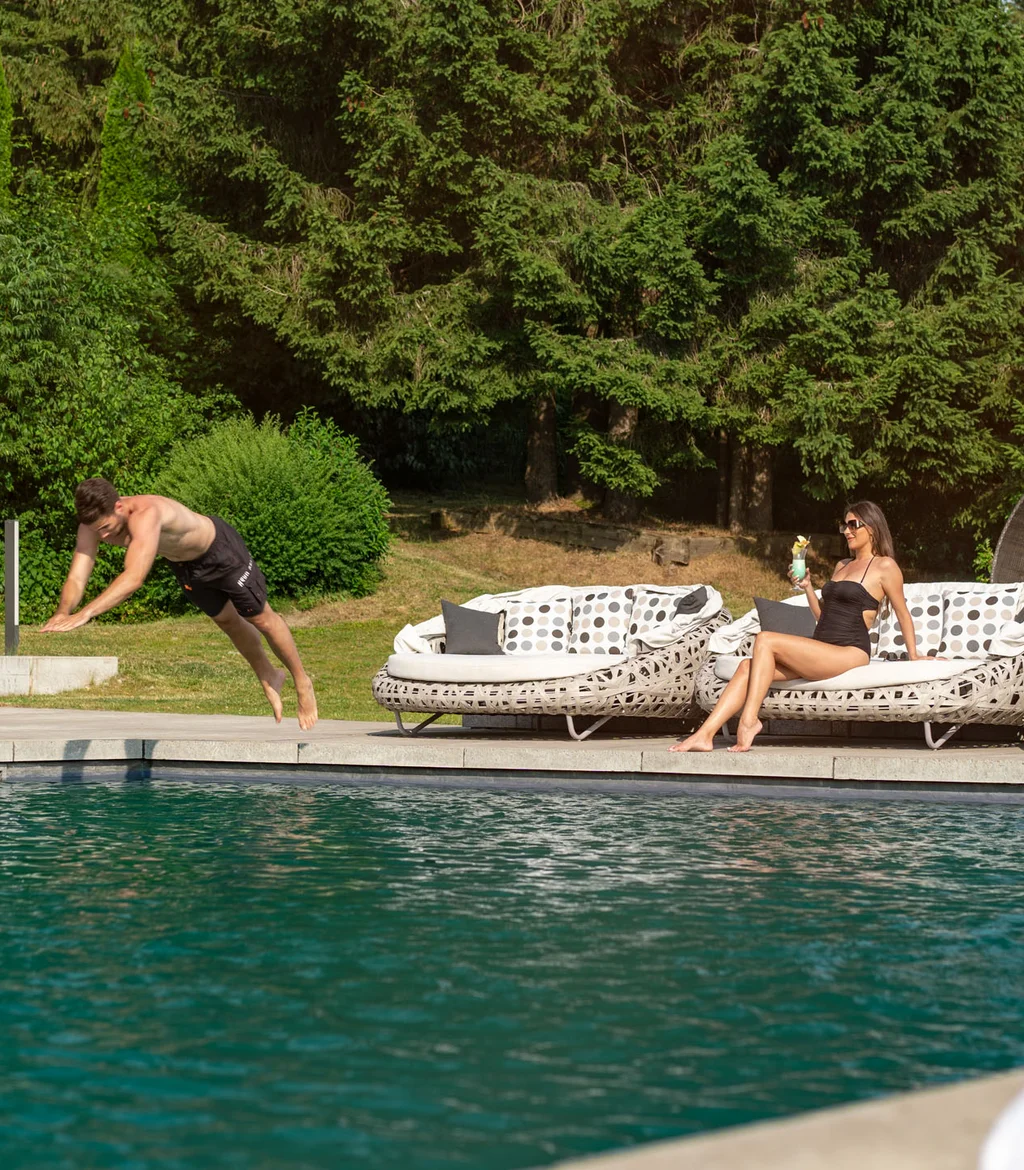 Mann springt ins Schwimmbecken, Frau sitzt entspannt mit Getränk auf Gartenliege