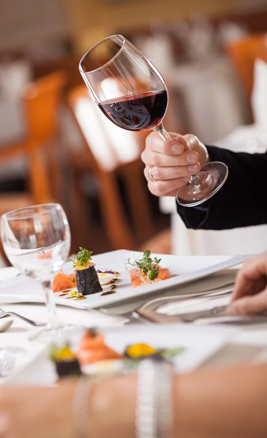 Die Adresse für Wellness in Bayern: Ihr Hotel in St. Englmar Hand hält Wein glas mit rotem Wein über einem gedeckten Tisch