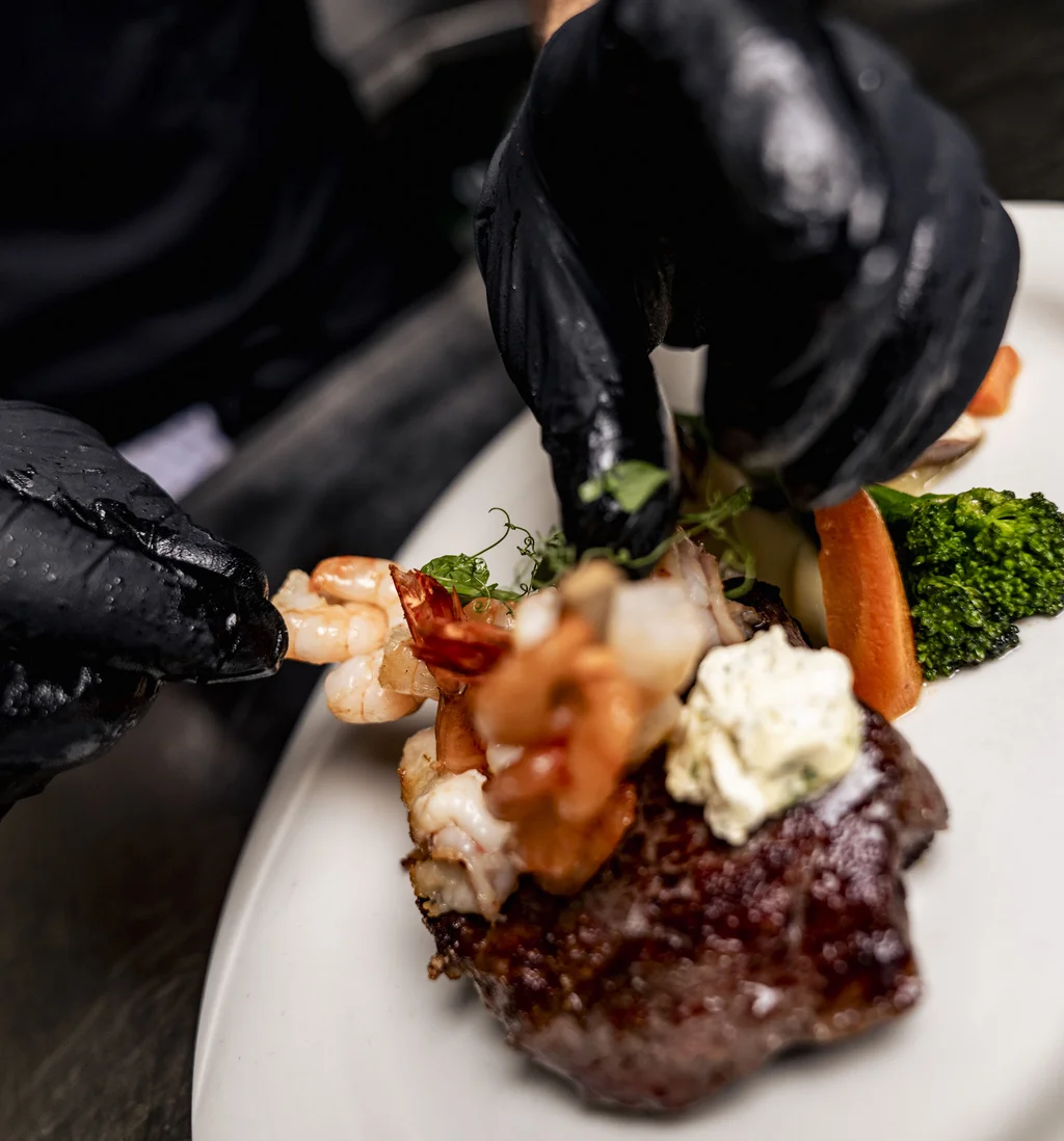 Chef legt mit Handschuhen Garnele auf Steak mit Gemüse