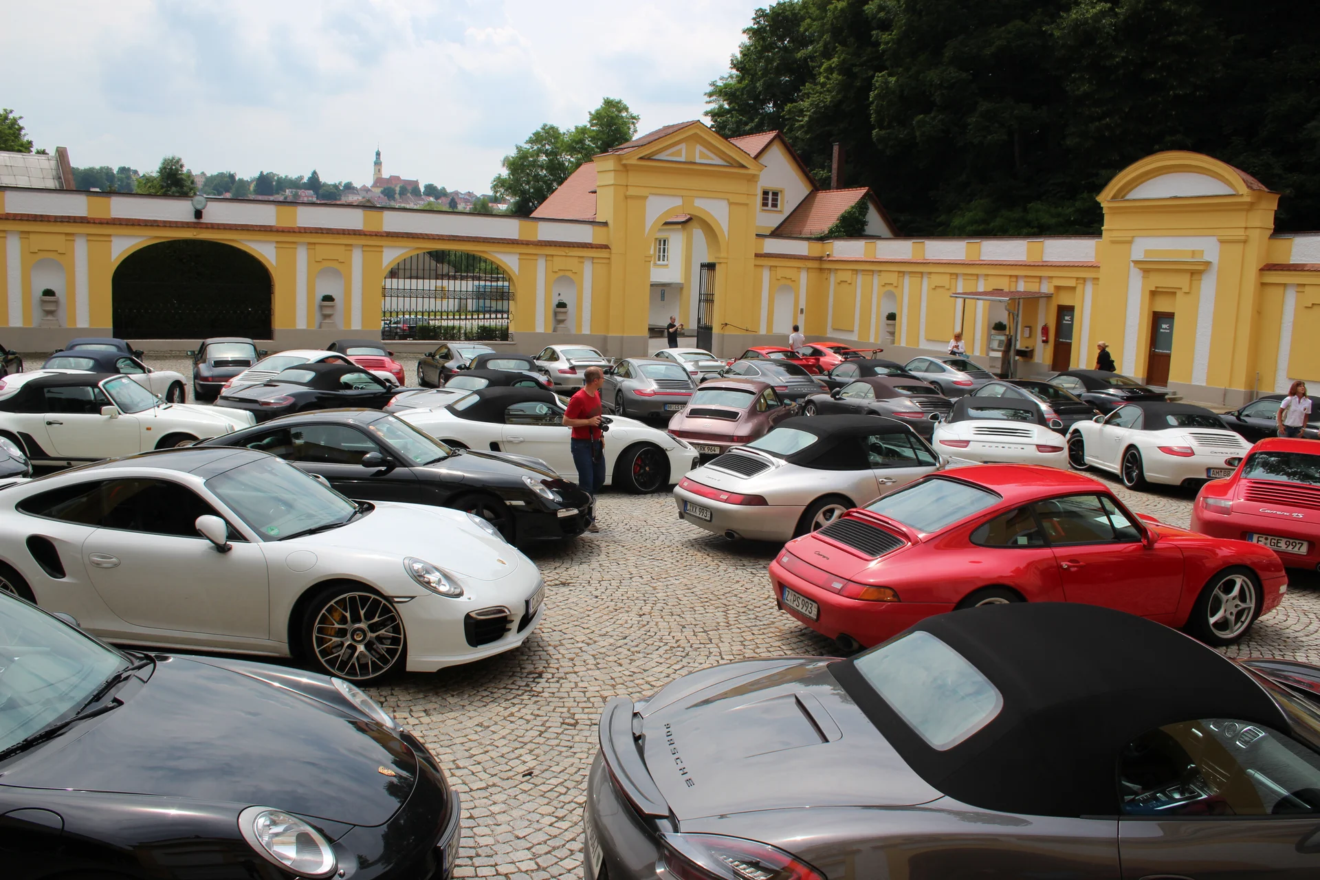 Viele Porsche-Sportwagen auf einem gepflasterten Parkplatz vor gelber historischer Mauer