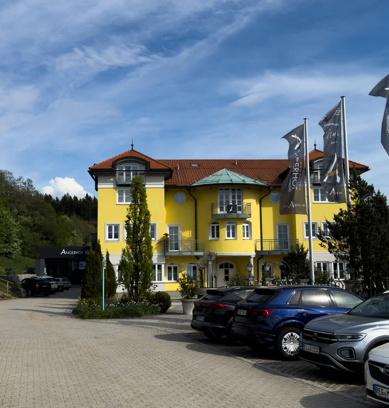 Die Adresse für Wellness in Bayern: Ihr Hotel in St. Englmar Gelbes Hotelgebäude mit rotem Dach und Flaggen vor blauem Himmel